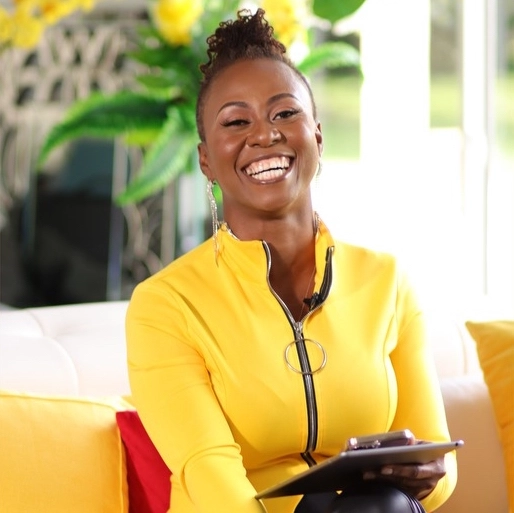 Smiling woman, named Chantale Milord, in a bright yellow jacket holding a tablet, sitting on a sofa, exuding warmth and positivity, ideal for coaching services or motivational speaking engagements.