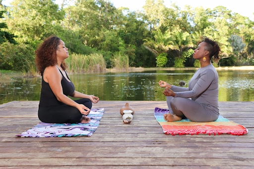 Two women sitting cross-legged on yoga mats practicing meditation outdoors by a peaceful lake, surrounded by lush greenery, promoting mindfulness and inner peace.