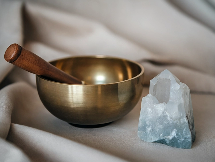 A brass Tibetan singing bowl with a wooden mallet placed beside a raw quartz crystal on soft beige fabric, symbolizing sound healing, meditation, and spiritual energy.